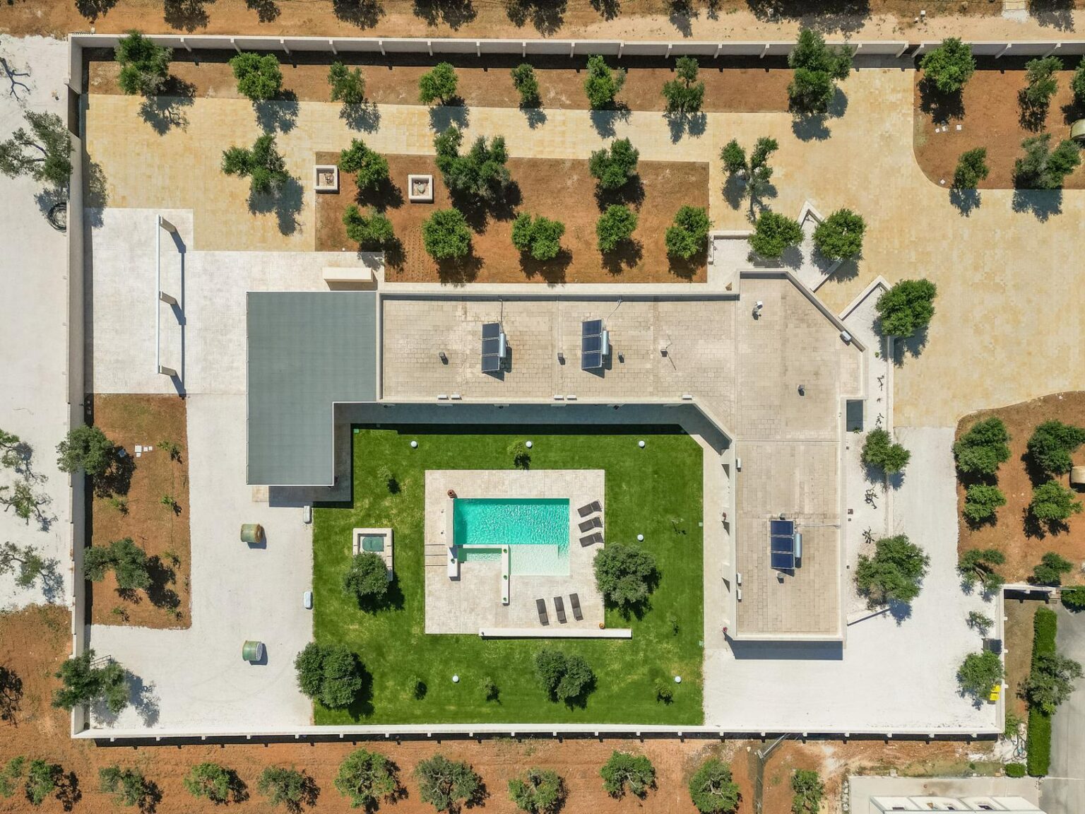 Aerial view of a modern residential property featuring a rectangular swimming pool surrounded by landscaped grass, sun loungers, and palm trees. The design includes a sleek patio area and structured landscaping, highlighting outdoor living spaces. | Only on SopranoVillas