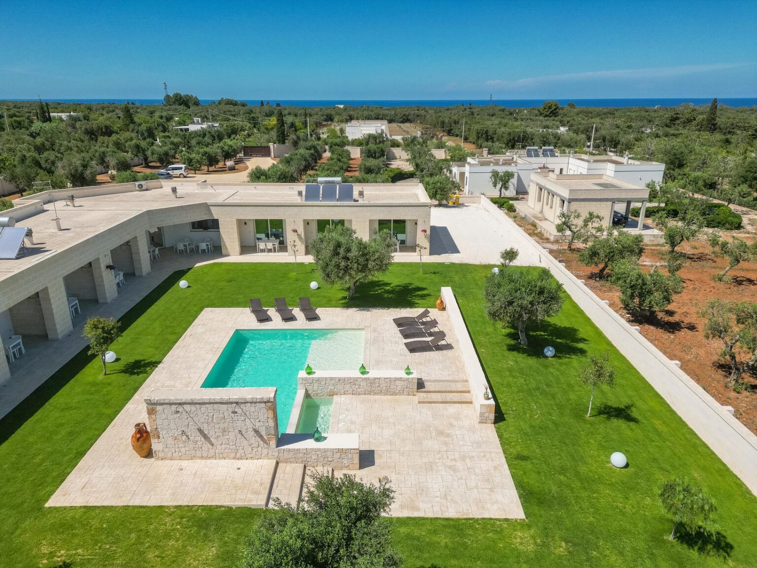 Aerial view of a modern villa with a swimming pool, surrounded by lush greenery and olive trees, overlooking the sea. The spacious outdoor area features lounge chairs and a well-maintained lawn, showcasing a luxurious and tranquil setting. | Only on SopranoVillas