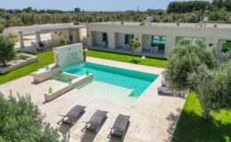 Aerial view of a modern villa with a turquoise swimming pool surrounded by lush greenery, sun loungers, and a serene outdoor space, ideal for relaxation and leisure.