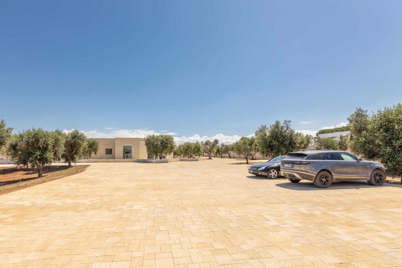 Spacious outdoor parking area featuring two cars, surrounded by olive trees and a clear blue sky, with a modern building in the background. | Only on SopranoVillas
