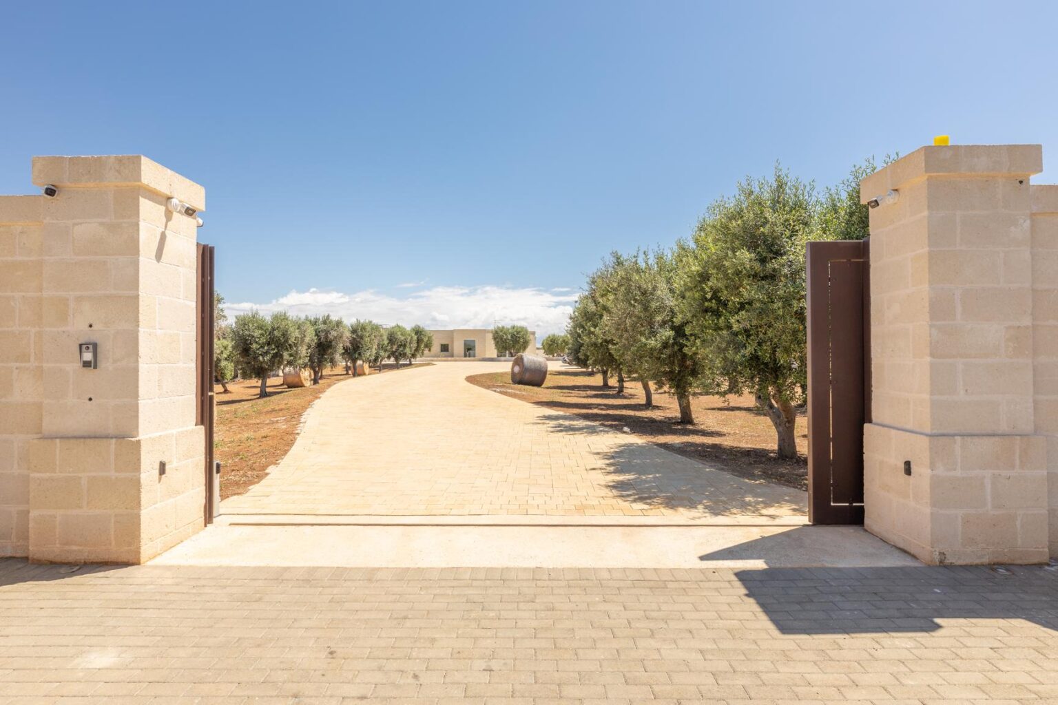 Rustic entrance with stone pillars and wooden gates leading to a scenic pathway lined with olive trees under a clear blue sky. | Only on SopranoVillas