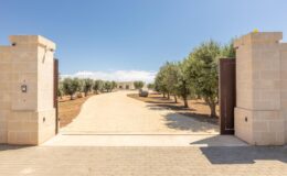 Rustic entrance with stone pillars and wooden gates leading to a scenic pathway lined with olive trees under a clear blue sky.