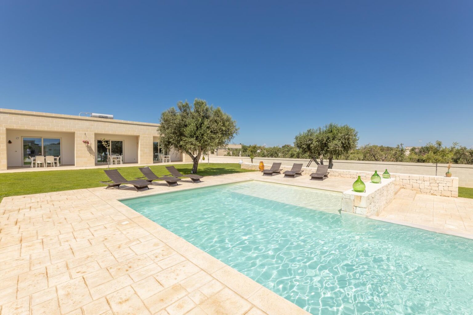 Modern outdoor swimming pool with lounge chairs, surrounded by green grass and trees under a clear blue sky. Ideal for relaxation and leisure activities. | Only on SopranoVillas