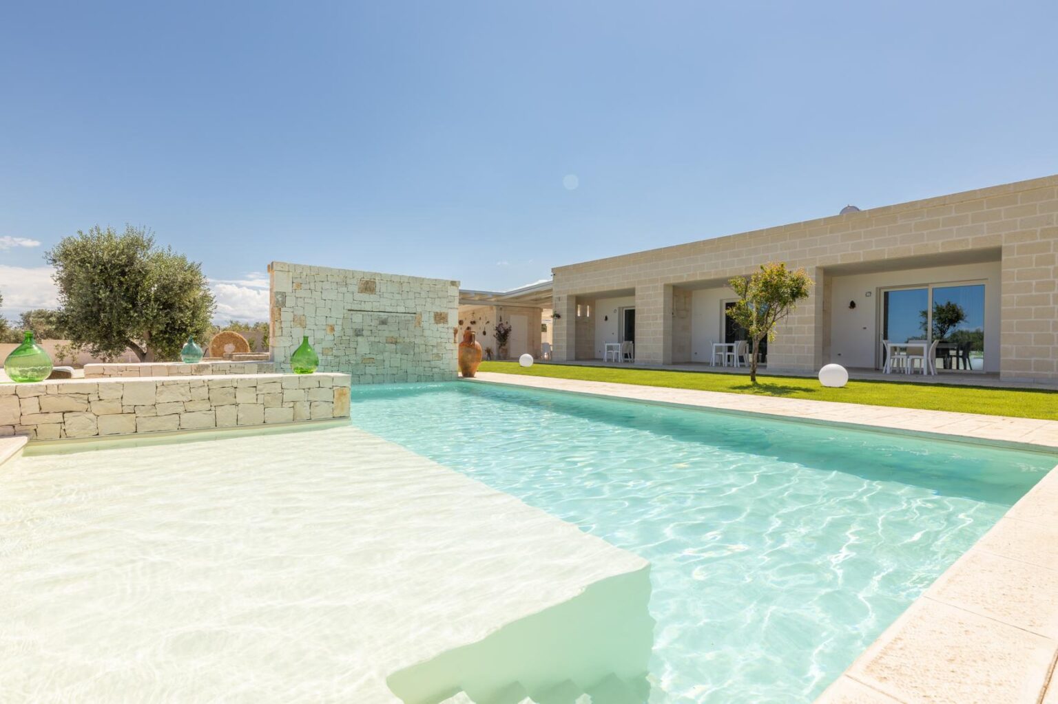 Modern villa with a swimming pool and landscaped garden under clear blue skies. The design features light-colored stone walls and a serene outdoor space, ideal for relaxation and leisure. | Only on SopranoVillas