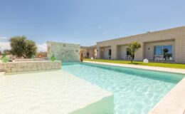 Modern villa with a swimming pool and landscaped garden under clear blue skies. The design features light-colored stone walls and a serene outdoor space, ideal for relaxation and leisure.