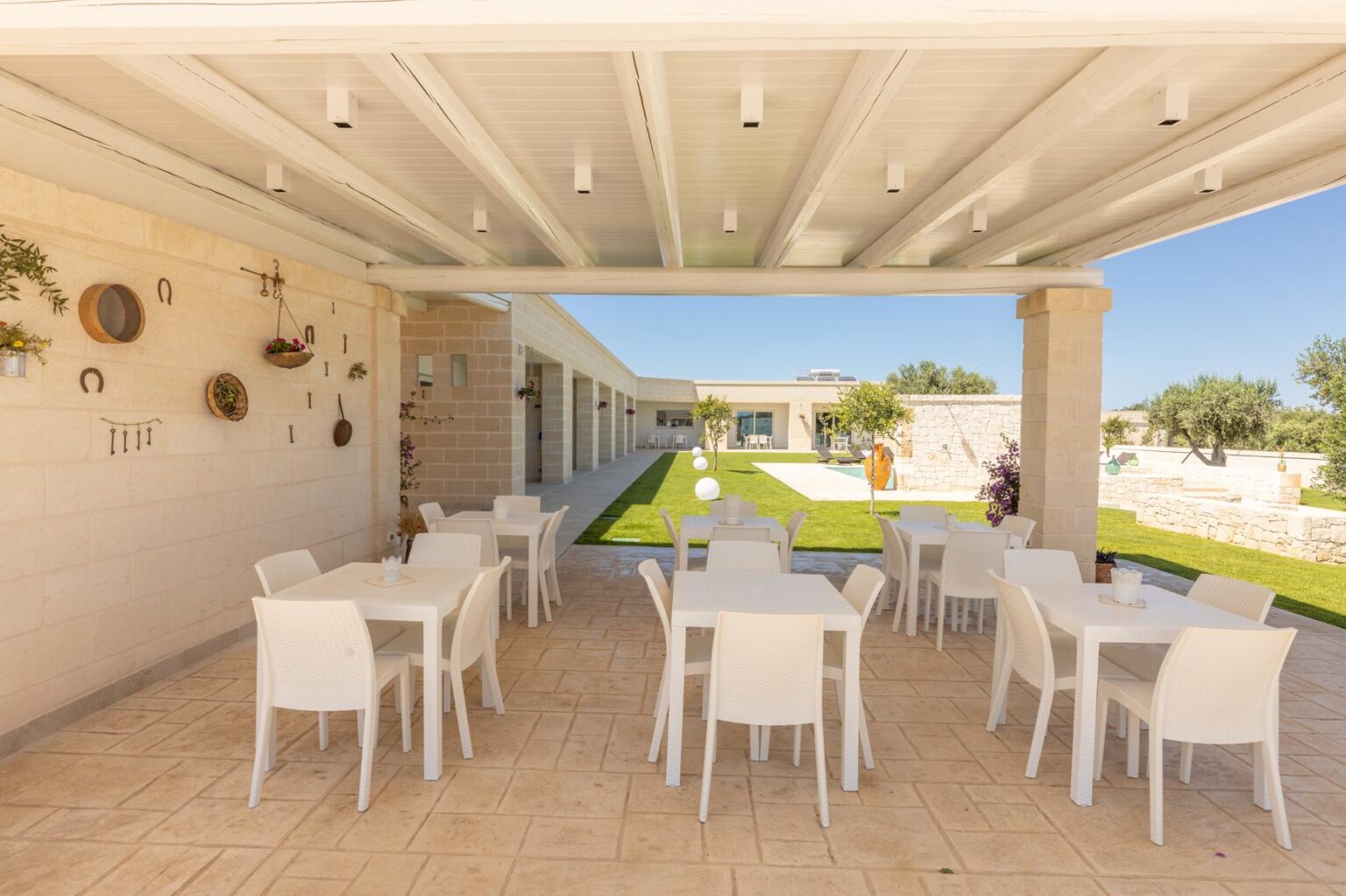 Outdoor dining area featuring white tables and chairs under a modern pergola, with a well-maintained lawn and decorative wall elements in the background, ideal for gatherings and events. | Only on SopranoVillas