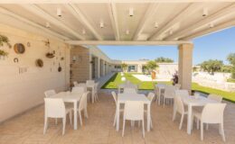 Outdoor dining area featuring white tables and chairs under a modern pergola, with a well-maintained lawn and decorative wall elements in the background, ideal for gatherings and events.