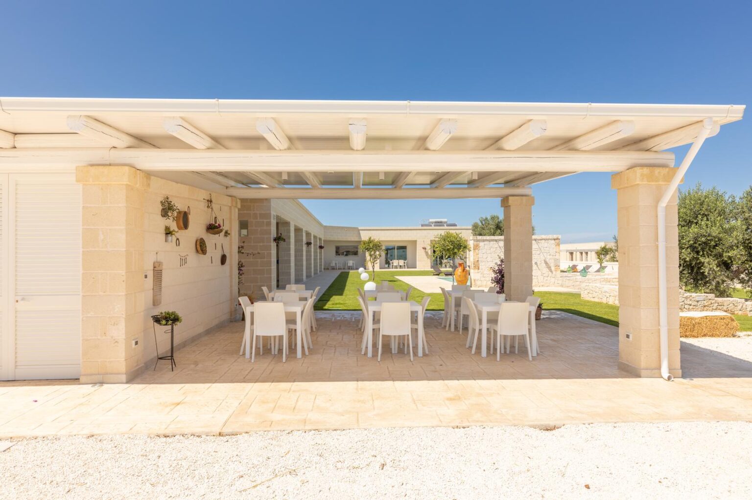 Modern outdoor dining area featuring a covered patio with white chairs and a scenic view of a landscaped garden. Ideal for gatherings and al fresco dining in a sunny environment. | Only on SopranoVillas