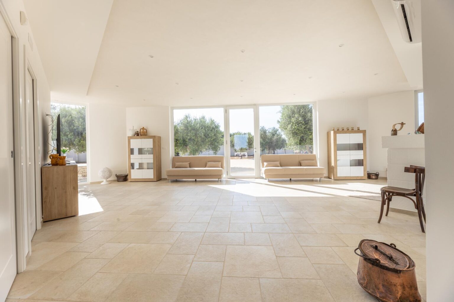 Spacious and bright modern living room with large windows, natural light, and minimalist decor, featuring beige sofas, wooden furniture, and a serene outdoor view. | Only on SopranoVillas