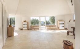 Spacious and bright modern living room with large windows, natural light, and minimalist decor, featuring beige sofas, wooden furniture, and a serene outdoor view.
