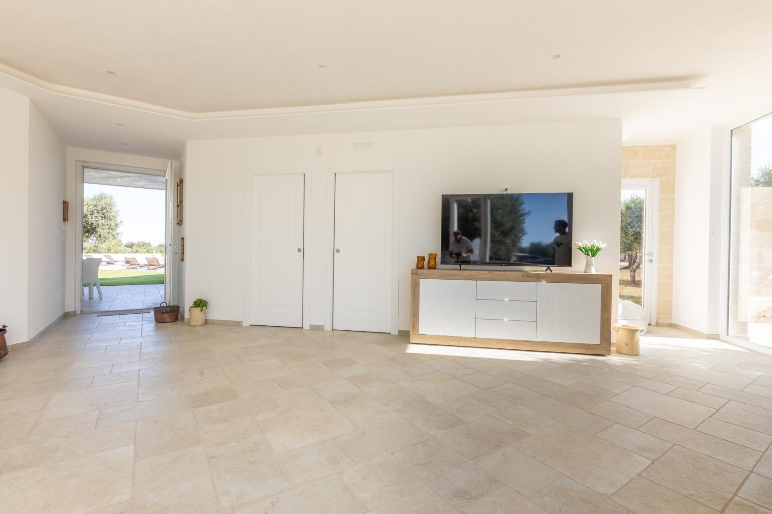 Modern living room featuring a minimalist design with light-colored walls, a large flat-screen TV mounted on a wooden console, and natural light streaming through sliding glass doors. The space has tiled flooring and a cozy atmosphere, ideal for relaxation and entertainment. | Only on SopranoVillas