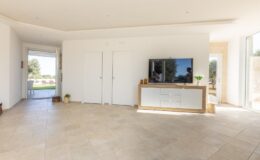 Modern living room featuring a minimalist design with light-colored walls, a large flat-screen TV mounted on a wooden console, and natural light streaming through sliding glass doors. The space has tiled flooring and a cozy atmosphere, ideal for relaxation and entertainment.