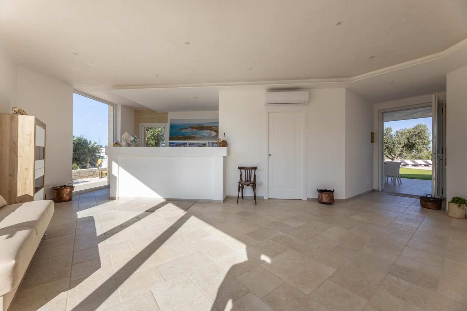 Bright and airy interior of a modern living space featuring a spacious layout, large windows, and natural light. The room includes a minimalist design with light-colored walls, tiled flooring, and a small wooden chair, creating a welcoming atmosphere. | Only on SopranoVillas
