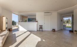 Bright and airy interior of a modern living space featuring a spacious layout, large windows, and natural light. The room includes a minimalist design with light-colored walls, tiled flooring, and a small wooden chair, creating a welcoming atmosphere.