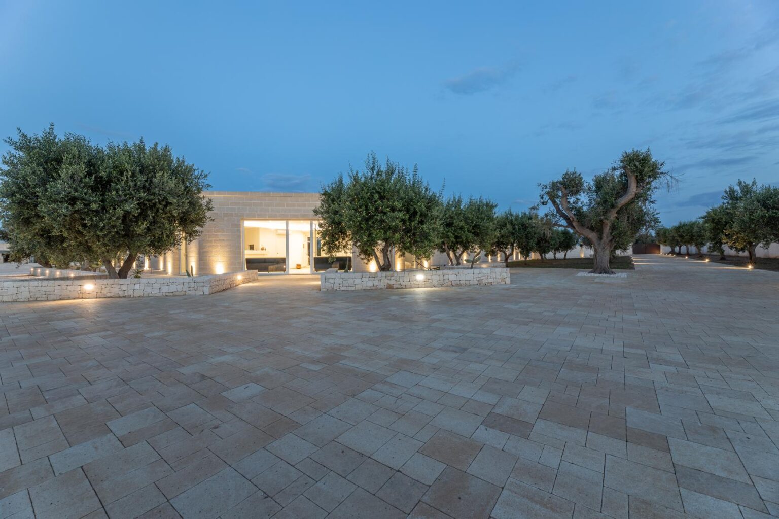 Modern architectural design of a house surrounded by olive trees, featuring a spacious stone-paved area, illuminated pathway, and a twilight sky. | Only on SopranoVillas