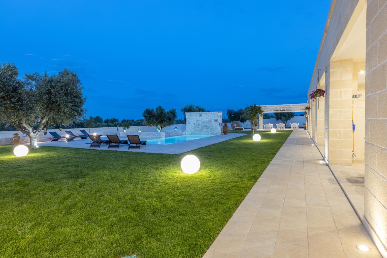 Modern villa exterior at dusk featuring a well-maintained lawn, a swimming pool with lounge chairs, and stylish outdoor lighting. | Only on SopranoVillas