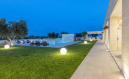 Modern villa exterior at dusk featuring a well-maintained lawn, a swimming pool with lounge chairs, and stylish outdoor lighting.