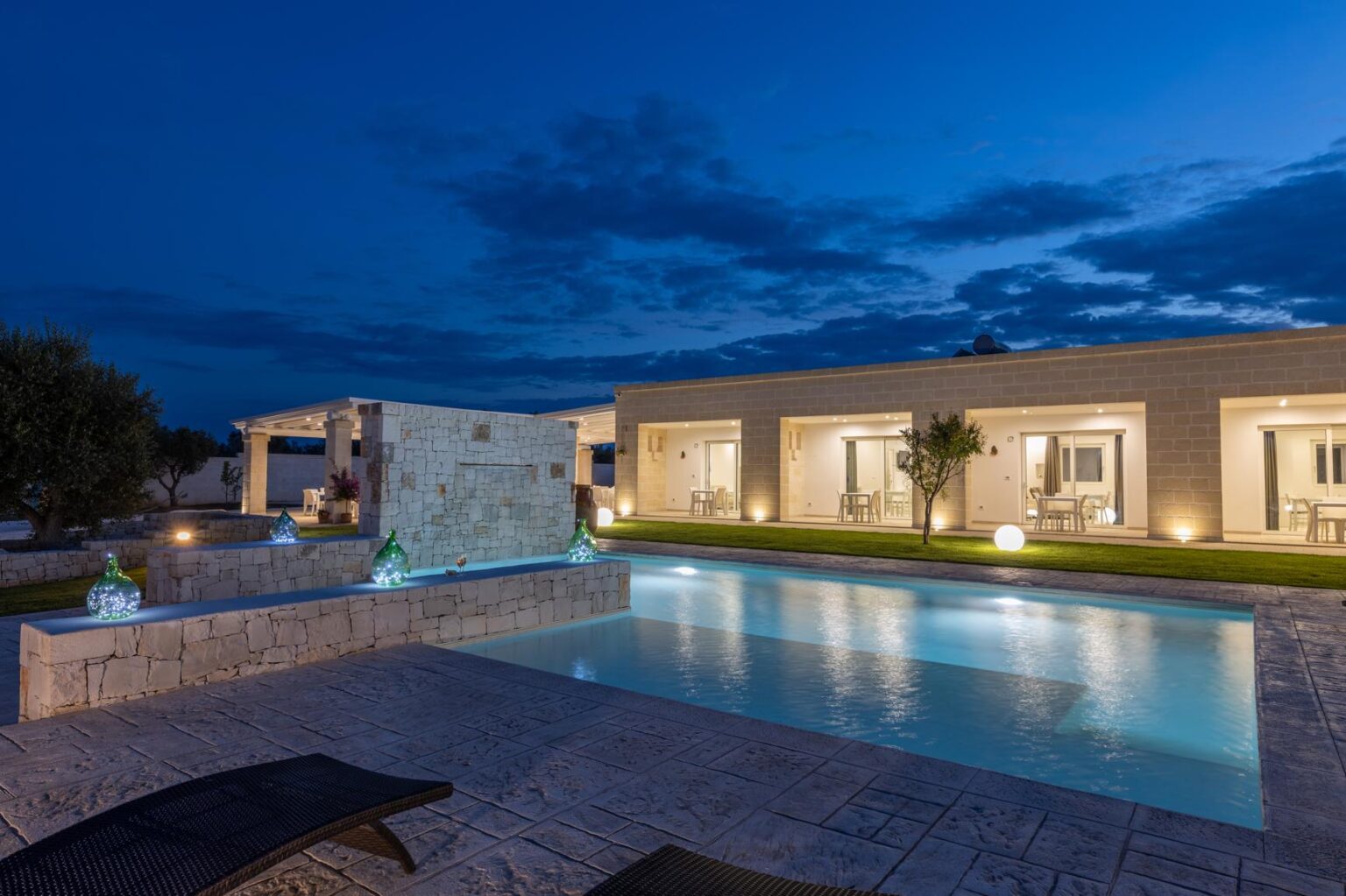 Modern villa with a swimming pool illuminated at night, featuring elegant stone architecture, landscaped gardens, and outdoor seating areas. Decorative glass sculptures enhance the serene atmosphere against a twilight sky. | Only on SopranoVillas