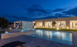 Modern villa with a swimming pool illuminated at night, featuring elegant stone architecture, landscaped gardens, and outdoor seating areas. Decorative glass sculptures enhance the serene atmosphere against a twilight sky.