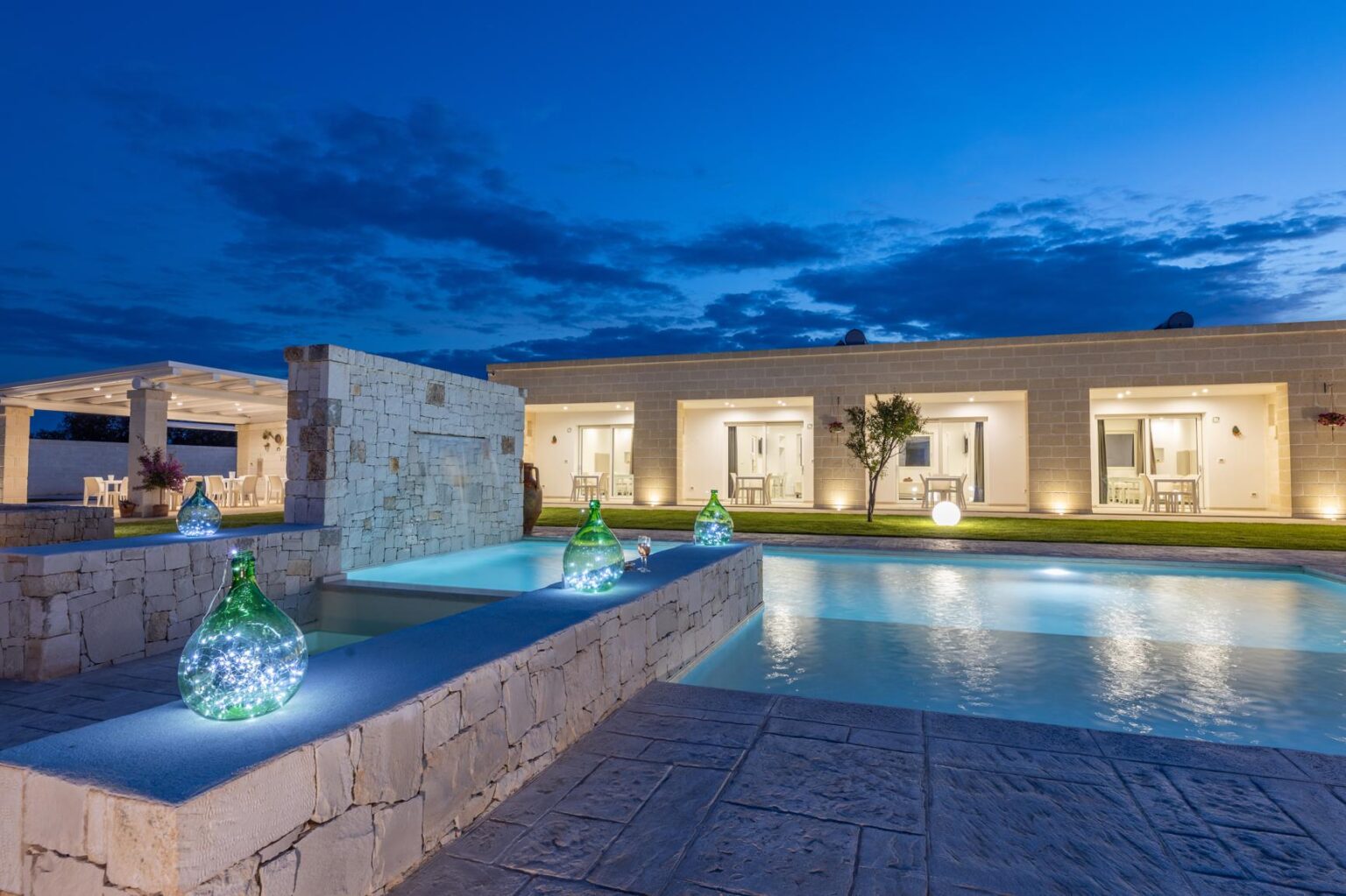 Modern luxury villa with a swimming pool and illuminated water features at dusk, showcasing stylish architecture and serene outdoor space. | Only on SopranoVillas