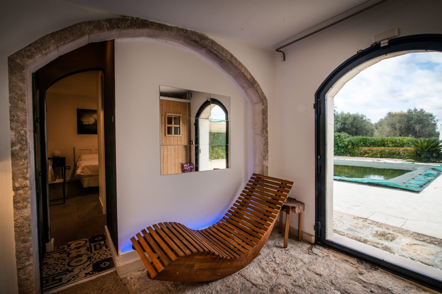 Cozy interior featuring a wooden lounge chair, stone walls, and large arched windows overlooking a serene outdoor pool and garden. The room showcases modern decor with a reflective mirror and rustic wooden accents, creating a relaxing atmosphere. | Only on SopranoVillas