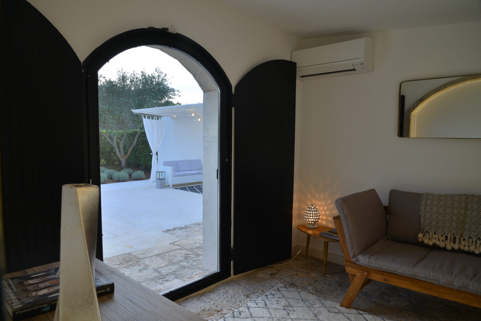 Interior view of a modern living space featuring an arched doorway with black shutters, a cozy seating area, and natural light illuminating the room. A patio is visible through the doorway, enhancing the indoor-outdoor connection. | Only on SopranoVillas