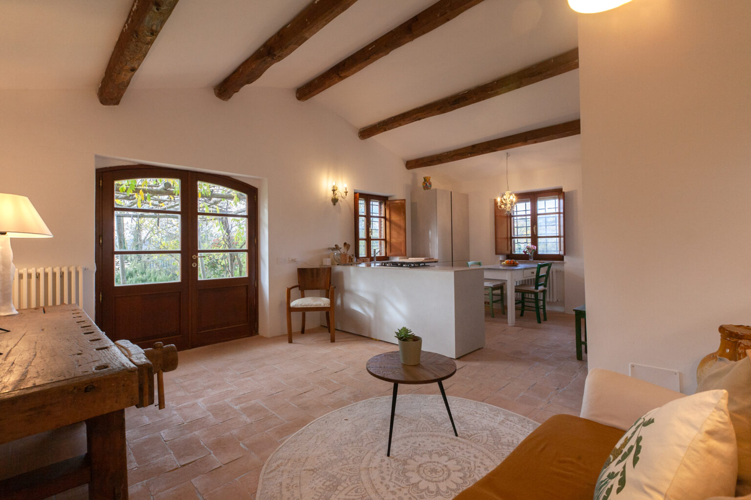 Cozy living room with wooden beams, natural light from large windows, and a rustic wooden table. A small round coffee table sits on a circular rug, creating a warm and inviting atmosphere in a stylish interior. | Only on SopranoVillas