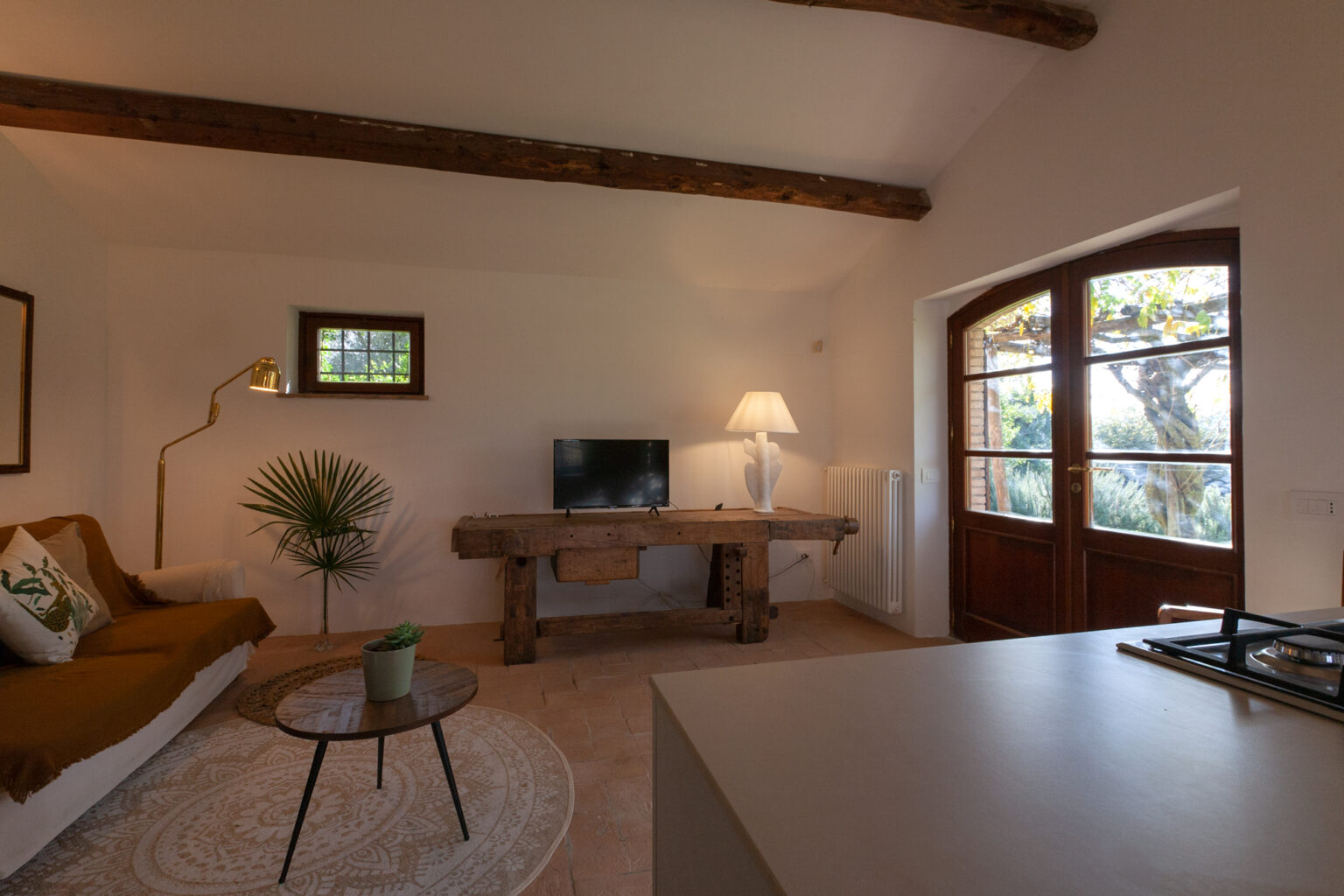 Cozy living room featuring a comfortable brown sofa, a rustic wooden coffee table, and a modern TV. Large windows with wooden frames allow natural light to fill the space, complemented by a decorative plant and a stylish lamp. Ideal for relaxation and entertainment in a serene setting. | Only on SopranoVillas