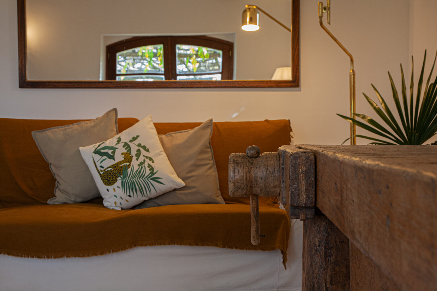 Cozy living room interior featuring a brown sofa with decorative pillows, a wooden table, and a stylish lamp. The space is enhanced by natural light coming through the windows and greenery in the background, creating a warm and inviting atmosphere. | Only on SopranoVillas