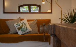 Cozy living room interior featuring a brown sofa with decorative pillows, a wooden table, and a stylish lamp. The space is enhanced by natural light coming through the windows and greenery in the background, creating a warm and inviting atmosphere.