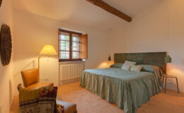 Cozy bedroom featuring a green bedspread, decorative pillows, a wooden headboard, and warm lighting from pendant lamps. A brown armchair with a colorful throw is positioned next to a window with wooden shutters, enhancing the inviting atmosphere. The room has stone flooring and a radiator, creating a comfortable and stylish retreat.