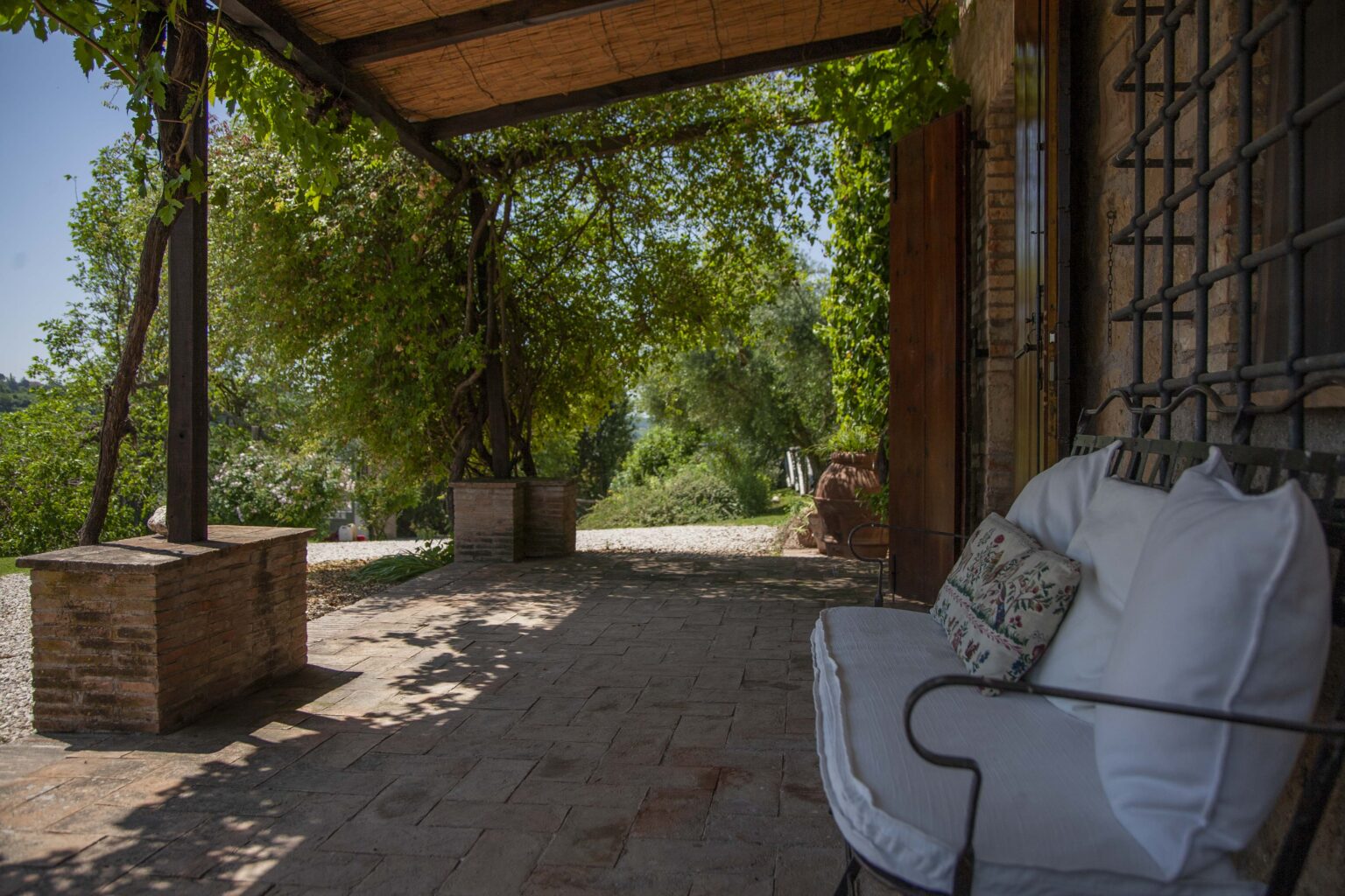 Charming outdoor seating area with a rustic stone floor, shaded by a wooden pergola adorned with lush greenery, offering a peaceful view of the surrounding landscape. Ideal for relaxation and enjoying nature. | Only on SopranoVillas