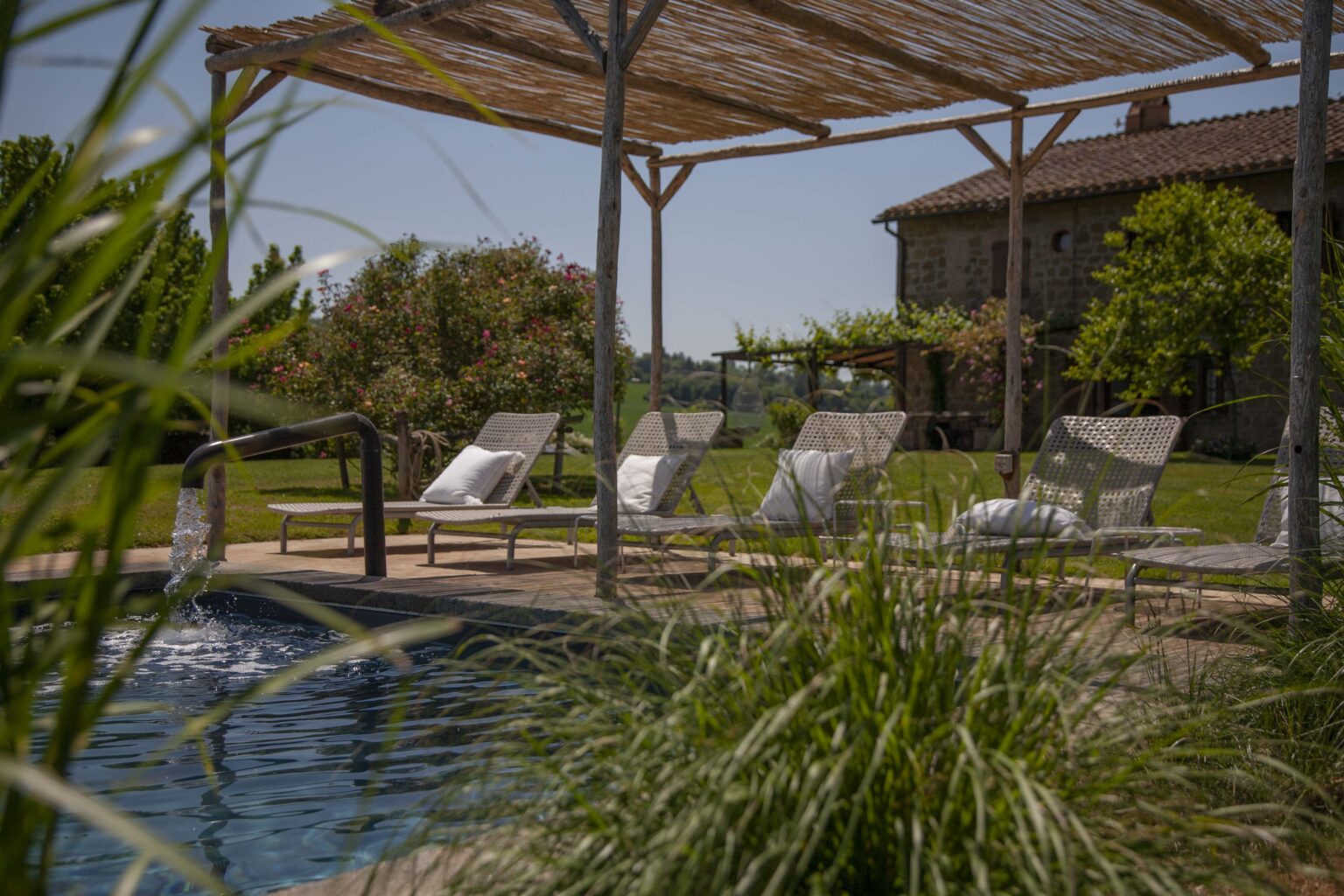 Serene outdoor pool area featuring comfortable lounge chairs under a thatched roof, surrounded by lush greenery and blooming flowers, with a rustic stone building in the background. Perfect for relaxation and enjoying nature. | Only on SopranoVillas