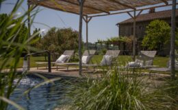 Serene outdoor pool area featuring comfortable lounge chairs under a thatched roof, surrounded by lush greenery and blooming flowers, with a rustic stone building in the background. Perfect for relaxation and enjoying nature.