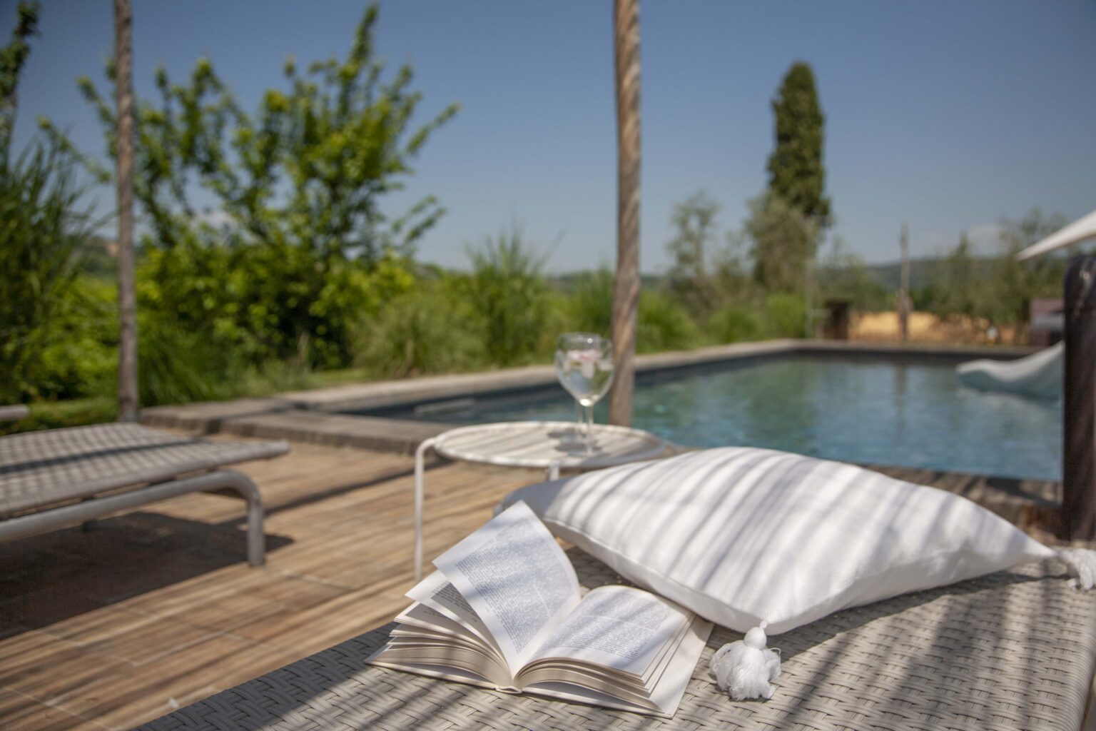 Relaxing outdoor lounge area with a pool, featuring comfortable chairs, decorative pillows, and a glass of drink, surrounded by greenery and a clear blue sky. | Only on SopranoVillas