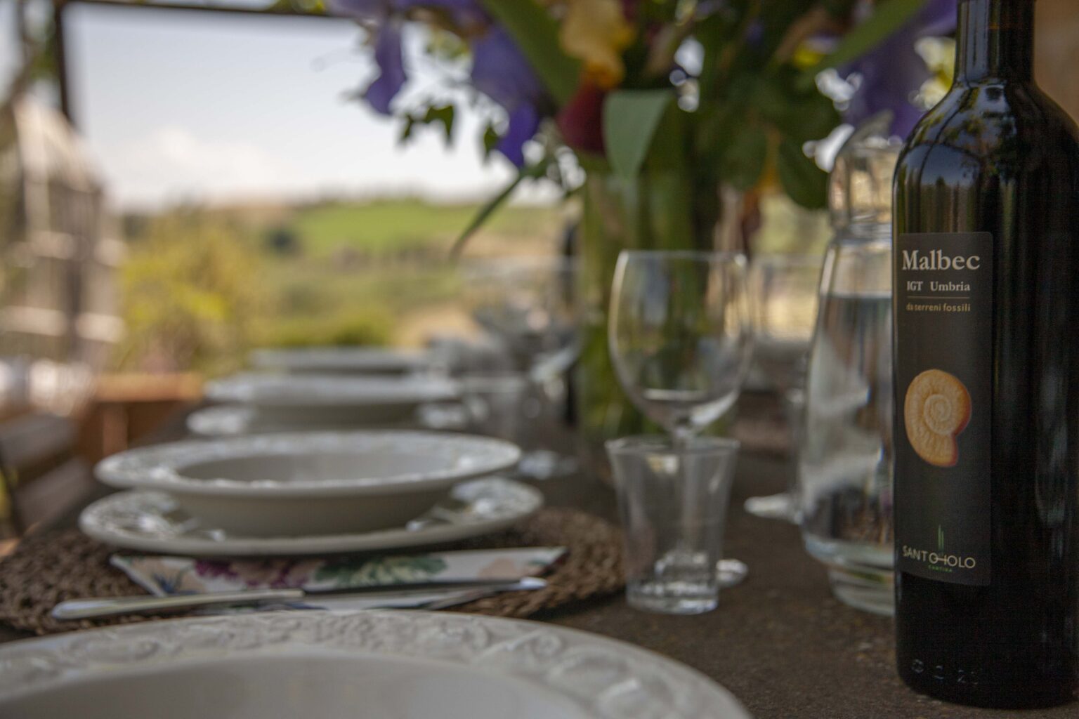 A beautifully set dining table outdoors featuring elegant white plates, wine glasses, and a bottle of Malbec wine from Umbria, surrounded by lush green hills and vibrant flowers in the background. | Only on SopranoVillas