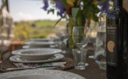 A beautifully set dining table outdoors featuring elegant white plates, wine glasses, and a bottle of Malbec wine from Umbria, surrounded by lush green hills and vibrant flowers in the background.