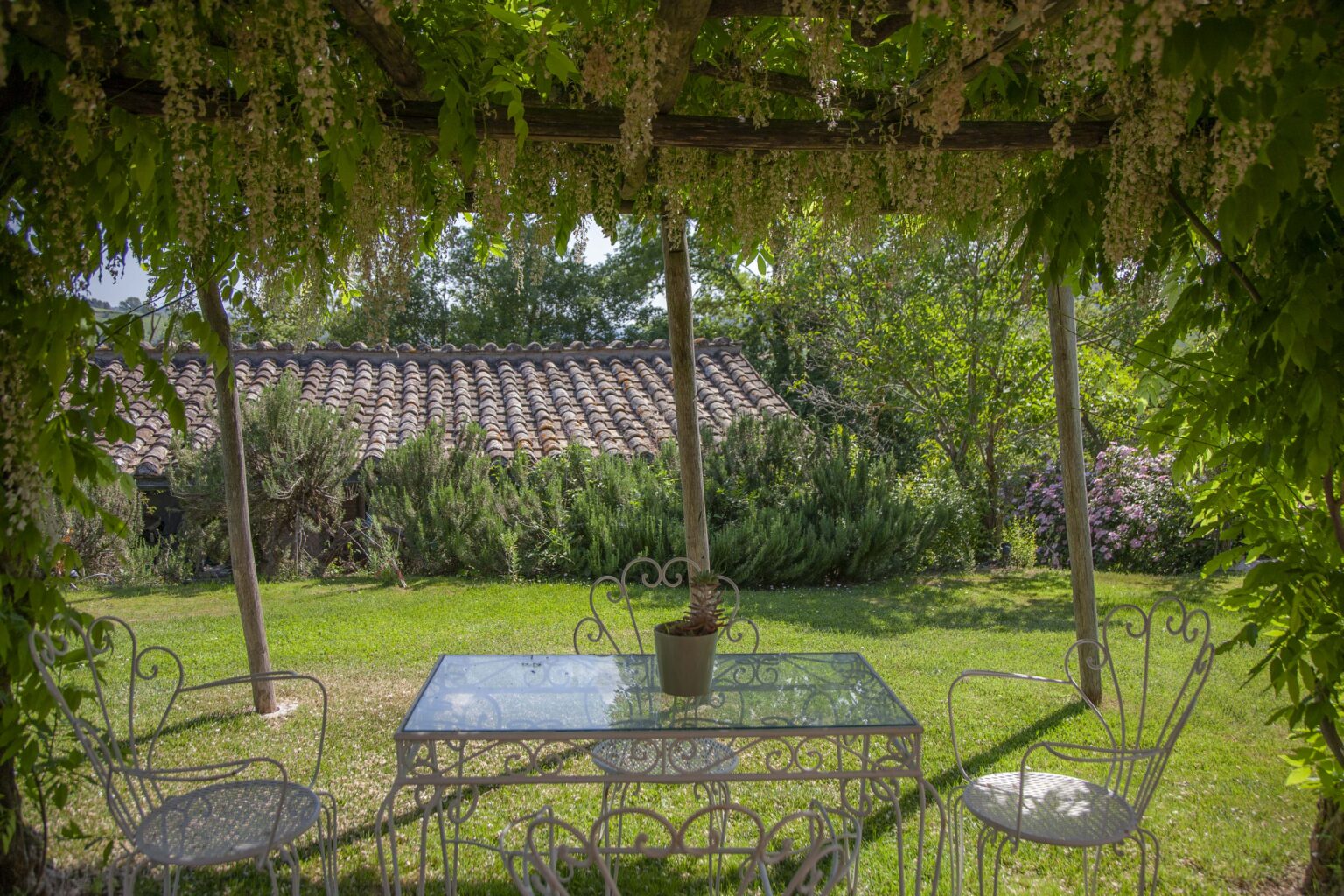Lush garden scene featuring a glass-top table and white wrought iron chairs under a pergola adorned with flowering vines, set against a backdrop of greenery and a rustic tiled roof. Ideal for outdoor dining or relaxation in a serene environment. | Only on SopranoVillas
