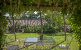 Lush garden scene featuring a glass-top table and white wrought iron chairs under a pergola adorned with flowering vines, set against a backdrop of greenery and a rustic tiled roof. Ideal for outdoor dining or relaxation in a serene environment.