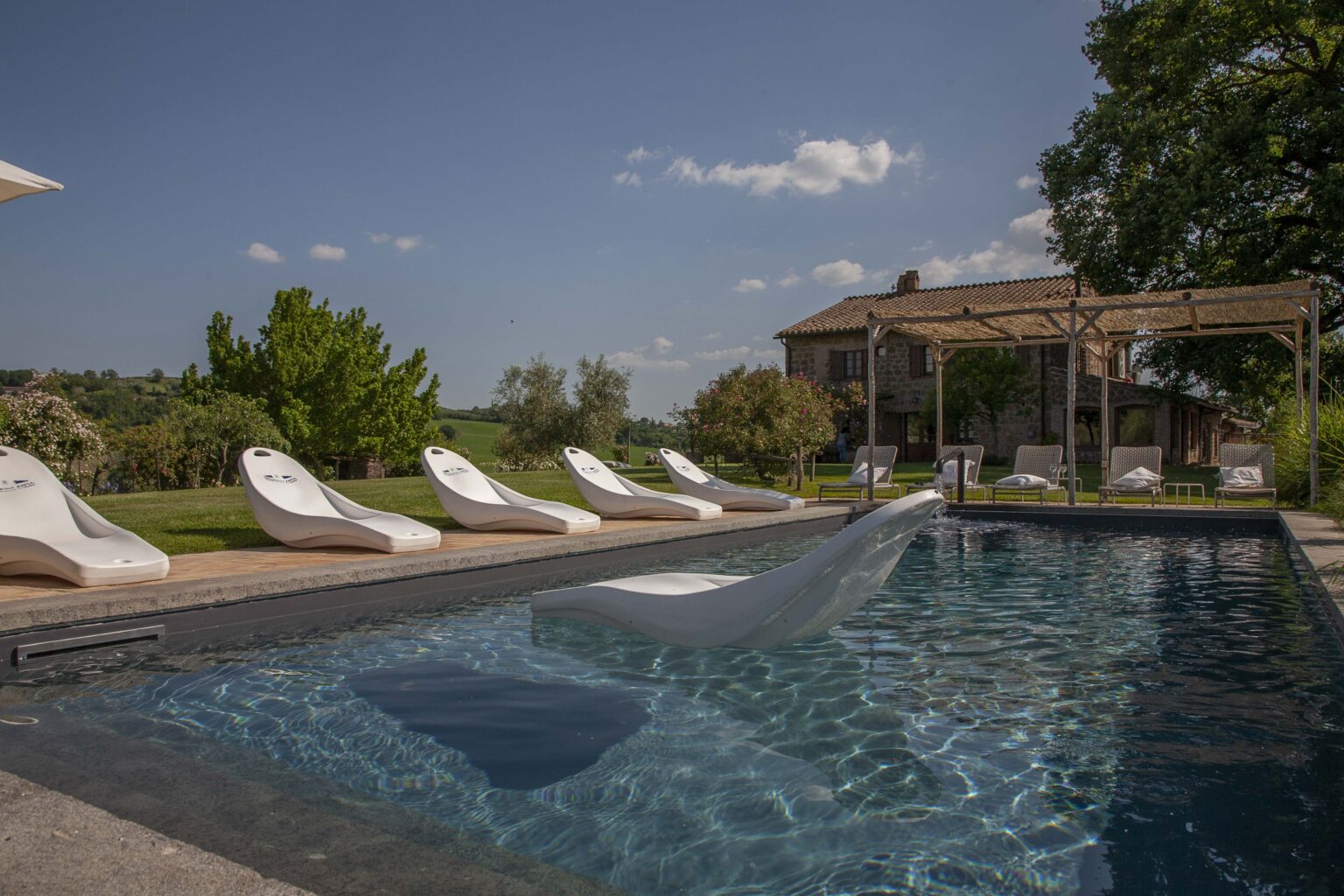 Luxurious outdoor swimming pool with modern loungers, surrounded by lush greenery and a picturesque villa in the background under a clear blue sky. | Only on SopranoVillas