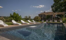 Luxurious outdoor swimming pool with modern loungers, surrounded by lush greenery and a picturesque villa in the background under a clear blue sky.