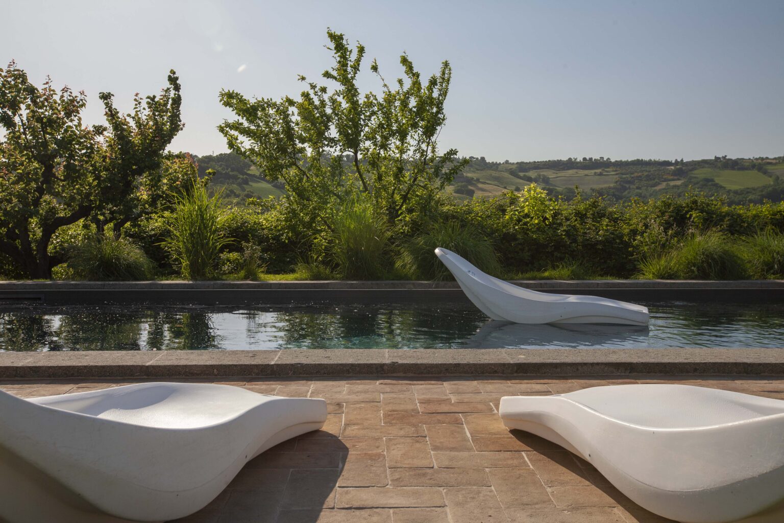 Modern outdoor lounge area featuring sleek white lounge chairs beside a tranquil swimming pool, surrounded by lush greenery and rolling hills under a clear blue sky. | Only on SopranoVillas