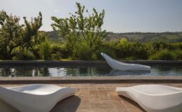 Modern outdoor lounge area featuring sleek white lounge chairs beside a tranquil swimming pool, surrounded by lush greenery and rolling hills under a clear blue sky.
