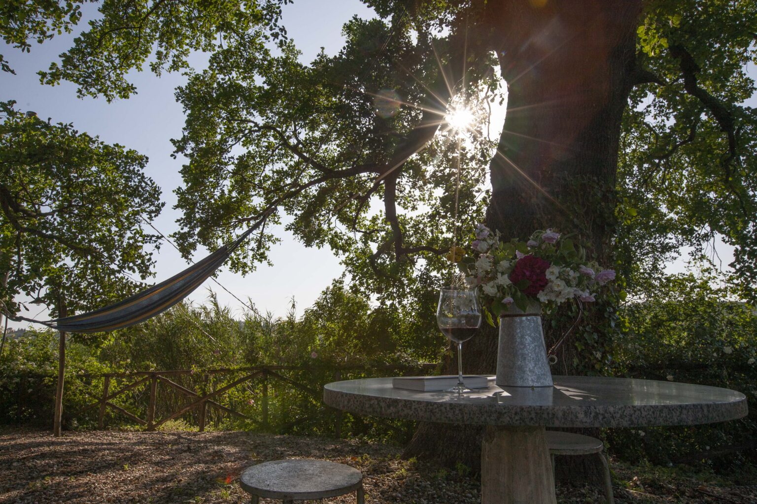 A tranquil outdoor setting featuring a hammock hanging between trees, a small table with a glass of wine and a vase of flowers, bathed in warm sunlight. | Only on SopranoVillas