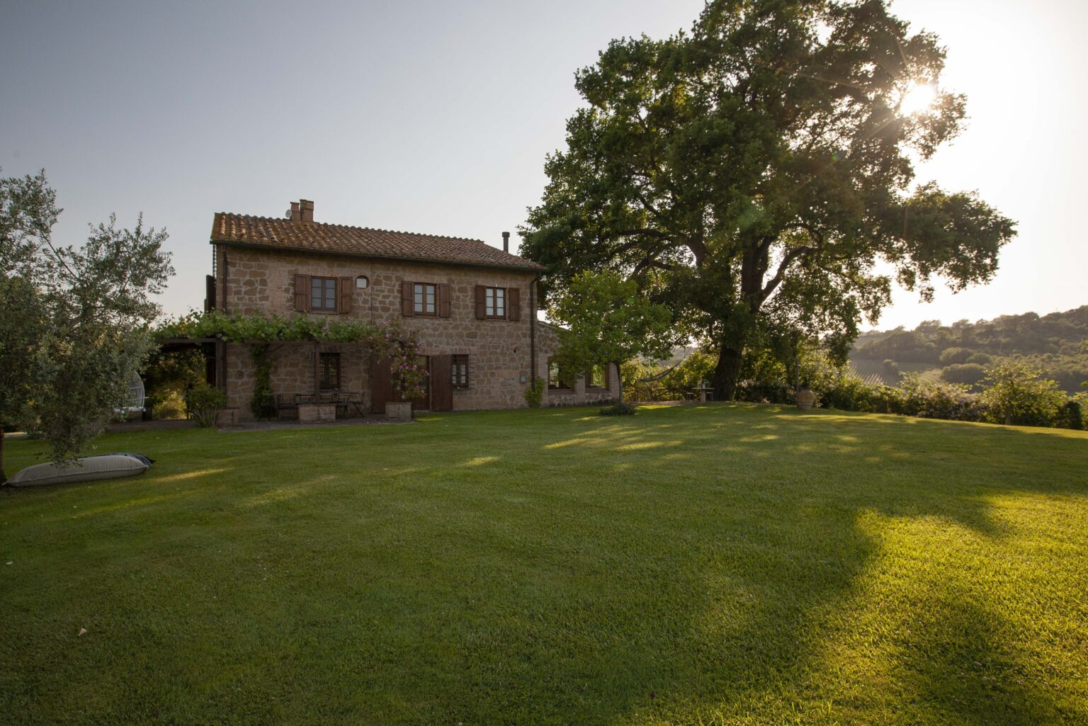 Charming rustic stone house surrounded by lush green lawn and trees, with a clear sky and sunlight casting shadows on the ground, ideal for a serene countryside retreat. | Only on SopranoVillas