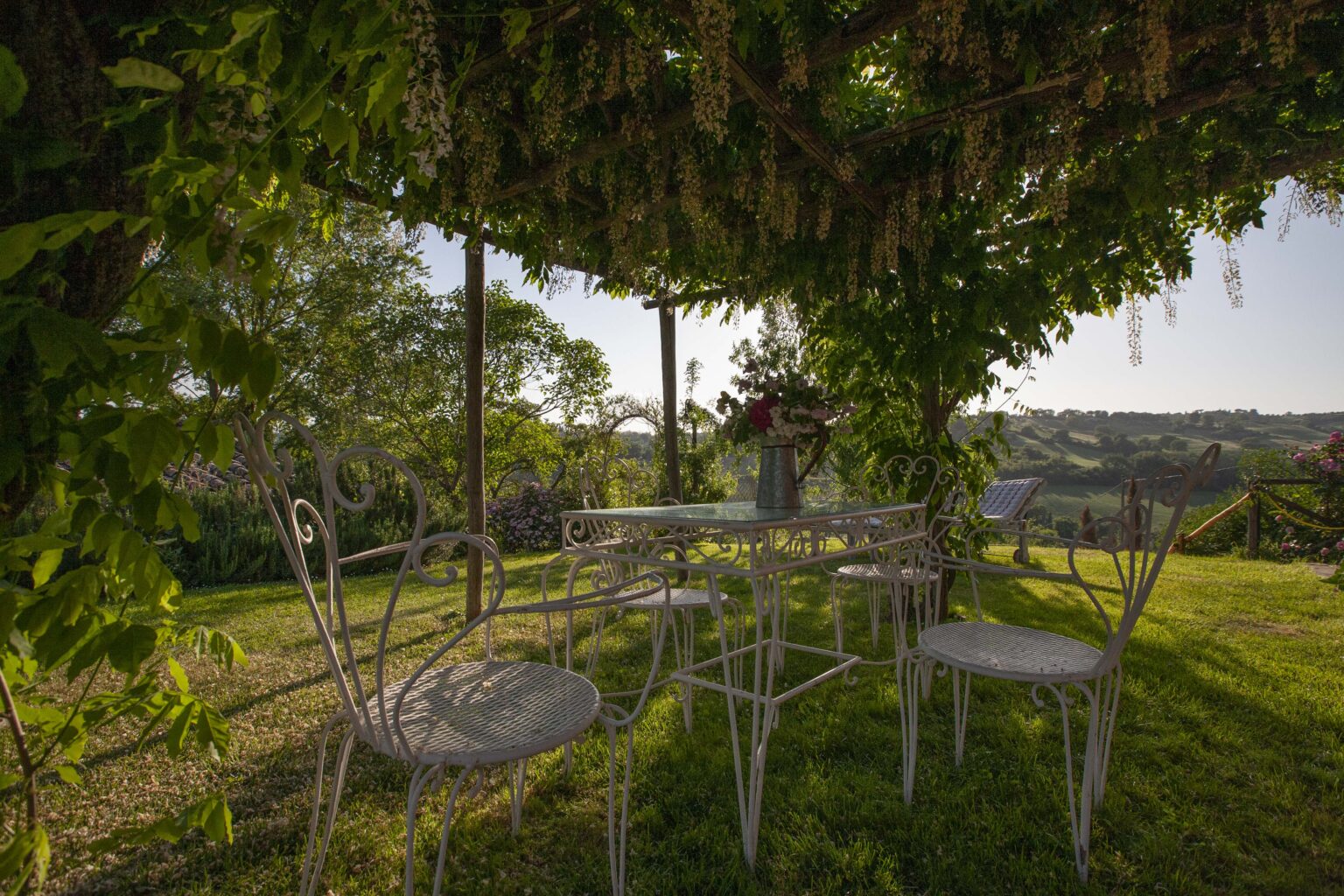 Charming outdoor seating area with a metal table and chairs under a lush pergola adorned with flowering vines, surrounded by a picturesque garden and rolling hills in the background. | Only on SopranoVillas