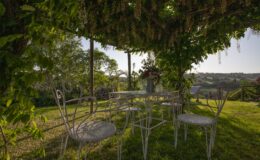 Charming outdoor seating area with a metal table and chairs under a lush pergola adorned with flowering vines, surrounded by a picturesque garden and rolling hills in the background.