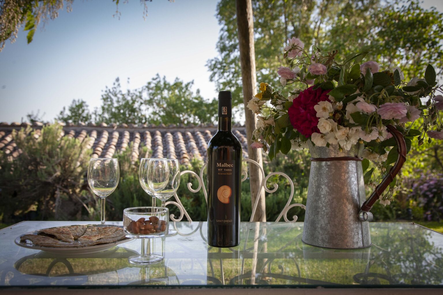 A serene outdoor setting featuring a bottle of wine, two wine glasses, a plate of snacks, and a vase of flowers on a glass table, with a backdrop of lush greenery and a rustic roof. Perfect for a relaxing wine tasting or outdoor gathering. | Only on SopranoVillas