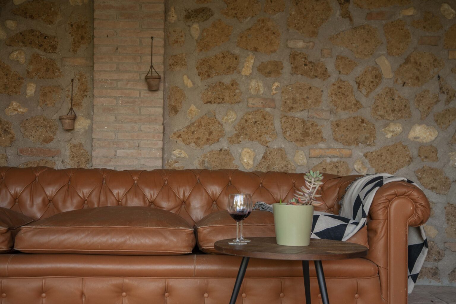 Chesterfield leather sofa in a cozy living room setting, accompanied by a small round table with a glass of red wine and a potted plant, against a rustic stone wall backdrop. | Only on SopranoVillas