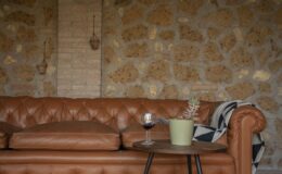 Chesterfield leather sofa in a cozy living room setting, accompanied by a small round table with a glass of red wine and a potted plant, against a rustic stone wall backdrop.
